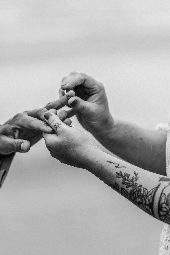 A tattooed bride slides a ring onto her grooms finger during their surfer elopement