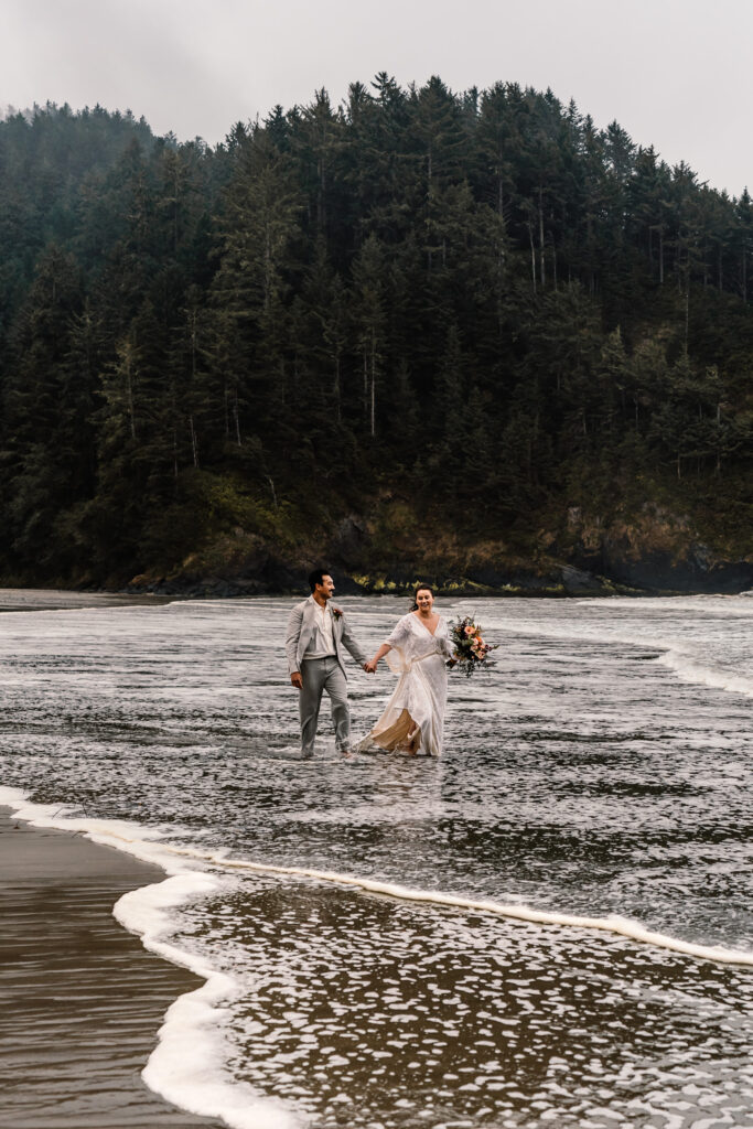 A bride and groom traipse through the tide as they explore a moody beach after their surfer wedding.