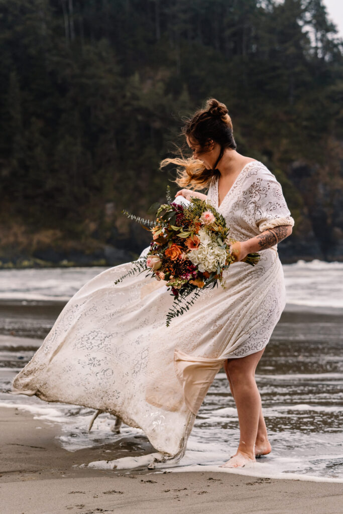 A bride walks on the beach during her surfer wedding as the wind billows her dress and whips her hair around wildly.