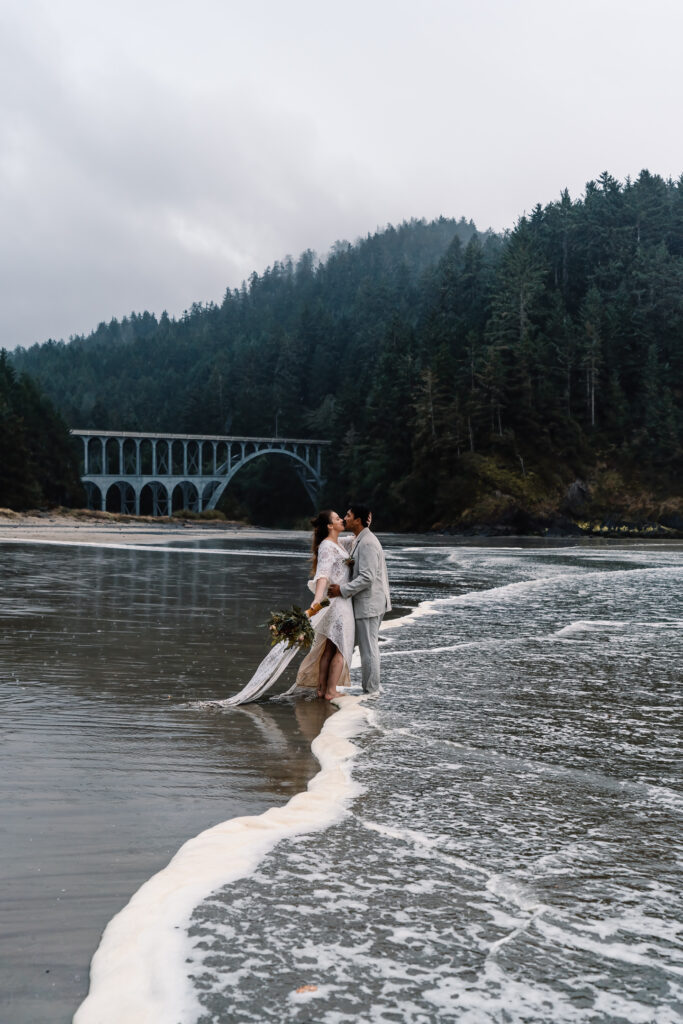 during their surfer wedding, a bride and groom kiss on the shore of a rocky beach, in front of a beautiful bridge. A wave of white sea foam rolls between them as they embrace.
