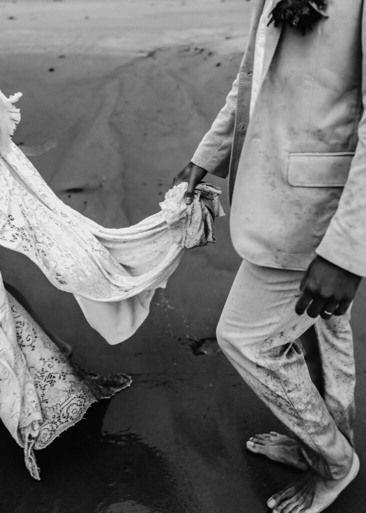 during their surfer wedding, a groom carries the sopping wet, and muddy train of his brides dress as they explore the beach.