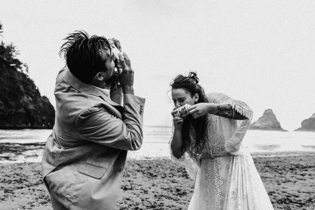 A black and white image of a couple shotgunning a beer to celebrate their elopement on the moody coast.