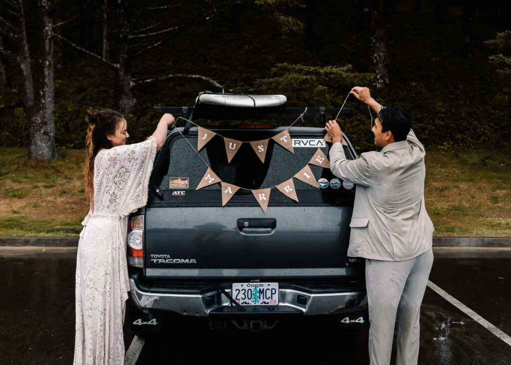 After their surfer wedding, a bride and groom hang a celebratory "just married" banner on the back of their car. 
