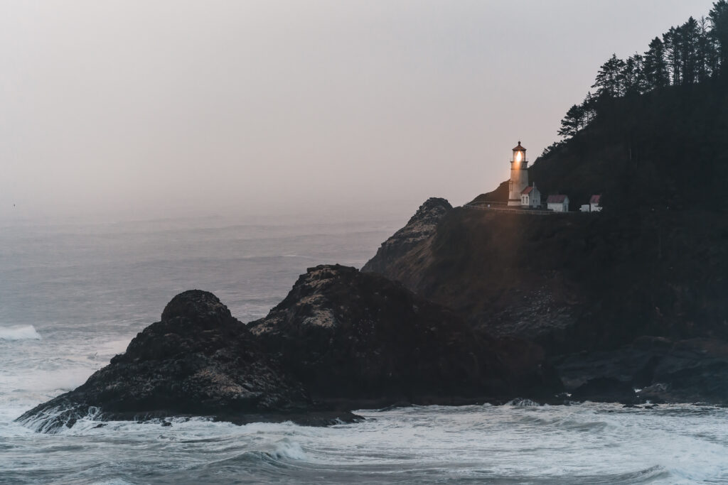 A landscape shot of a lighthouse on a seaside cliff. Its line shines brightly through the moody storm that has rolled in