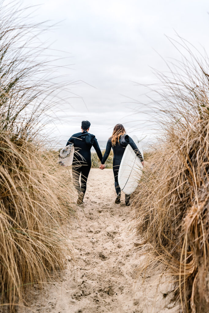 during their surfer wedding, a bride and groom walk through a tunnel of sea grass with their boards. They wear their wetsuits as they prepare for a brisk surf session