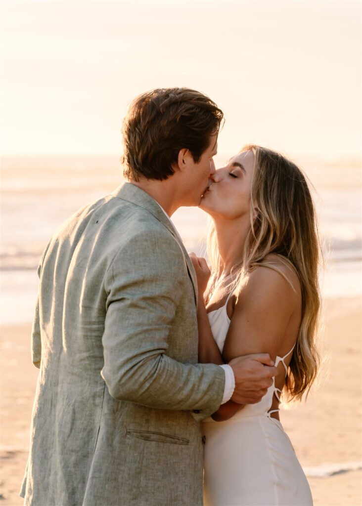 during their coastal elopement, a bride and groom embrace and kiss as the sun flares gold behind them. 