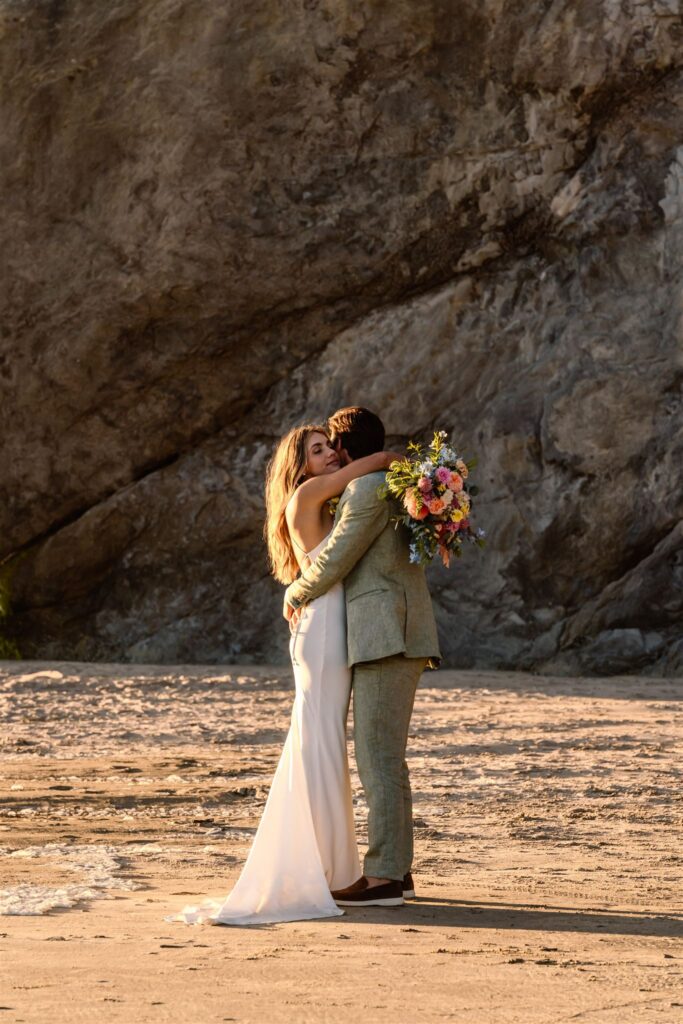 during their coastal elopement a bride and groom embrace in front of a  large sea rock. she flings her bouquet over his shoulder as she embraces him. 