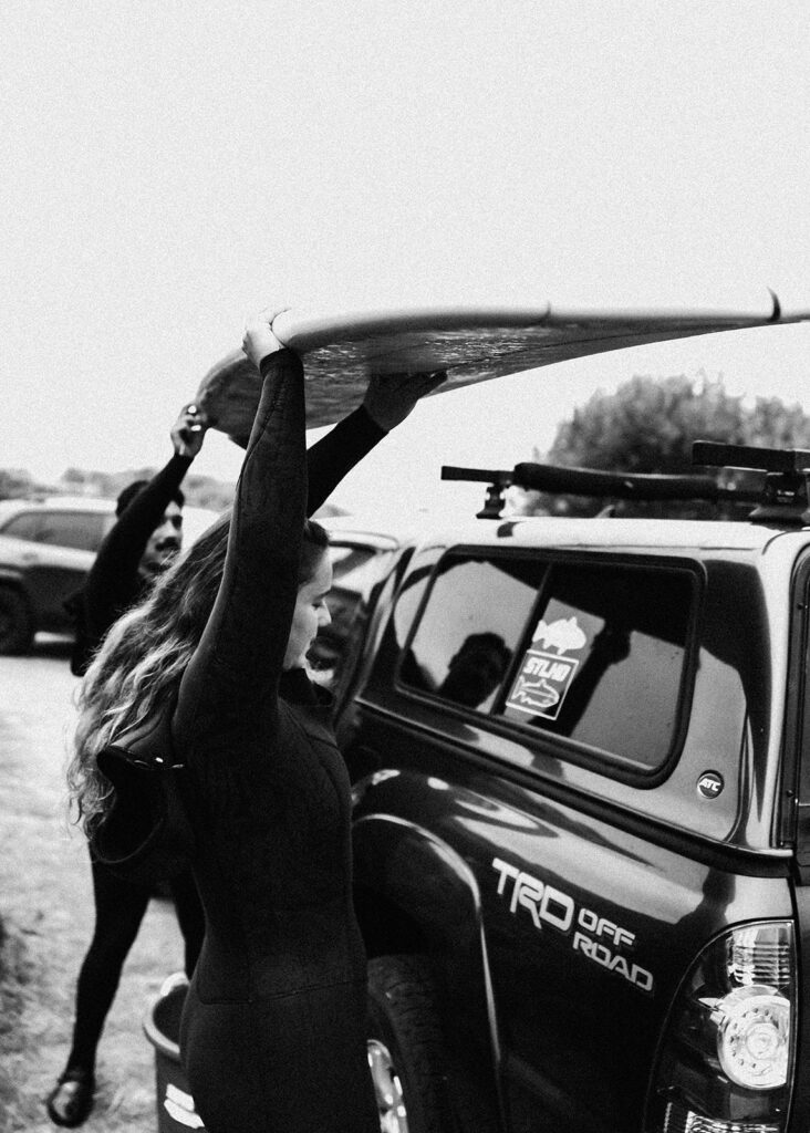 during their surfer wedding, a bride and groom dismount their boards from the roof rack of their car. They both wear wet suits as they prepare for a quick adventure. 