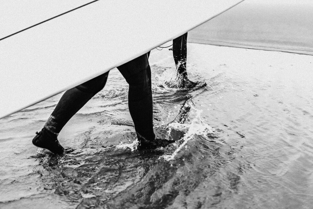 during their surfer wedding, a couple in their wetsuits walk through puddles on the beach. Their steps cause the puddles to splash as their boards frame their feet, becoming the focus of this story-telling image.
