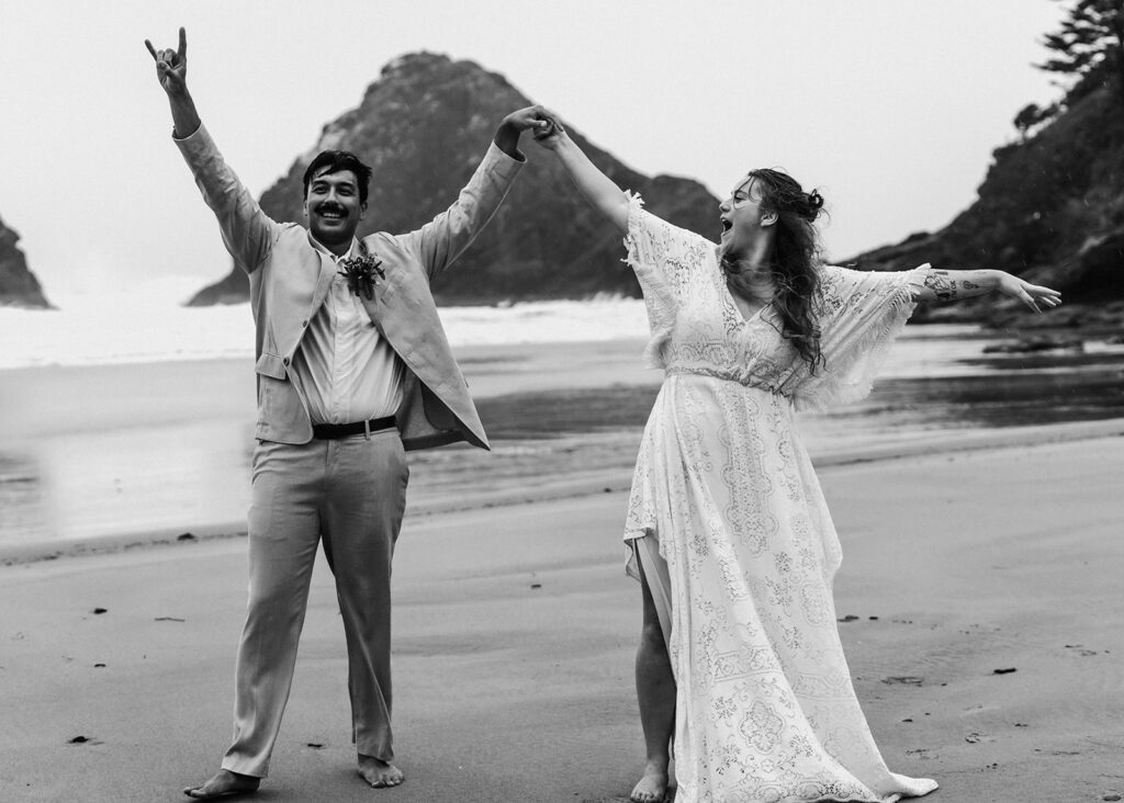 During their surfer wedding, a bride and groom cheer on a rocky beach. They raise their hands in exclamation and cheer excitedly after exchanging vows on the rocky beach. Rain pours down on them.