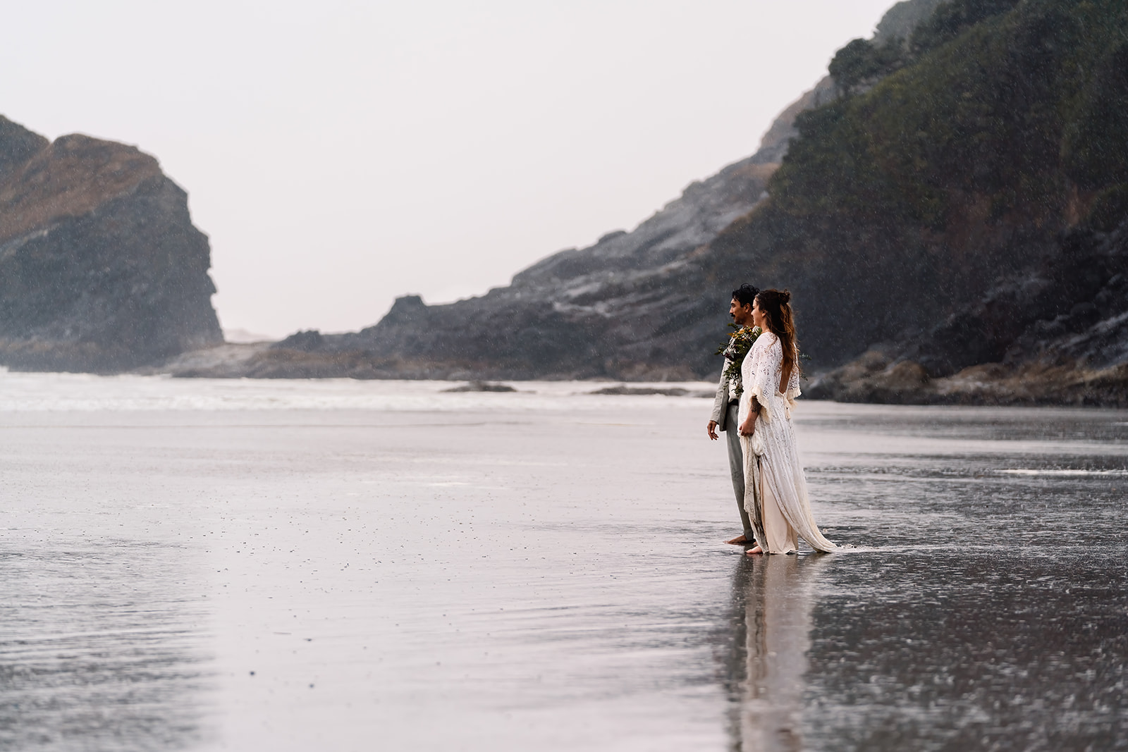 During their surfer wedding, a couple gazes out towards the moody ocean. The rain can be seen pouring around them as they stand in awe of the sea.