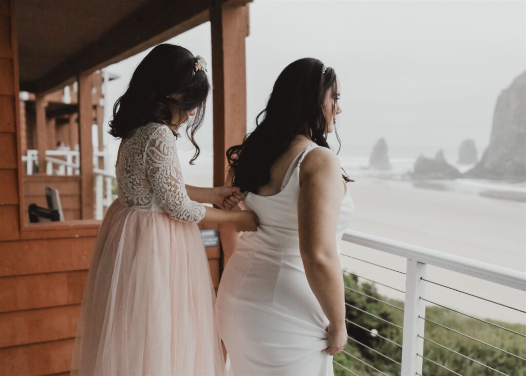 A young girl in a pink and white dress helps her mother prepare for her Pacific coast wedding. Her mother gazes towards the beach as she zips her dress. 