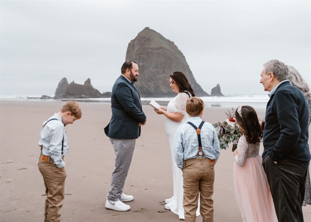 a bride and groom exchange vows in front of a haystack rock on a gloomy beach. They are surrounded by their family during their pacific coast wedding 