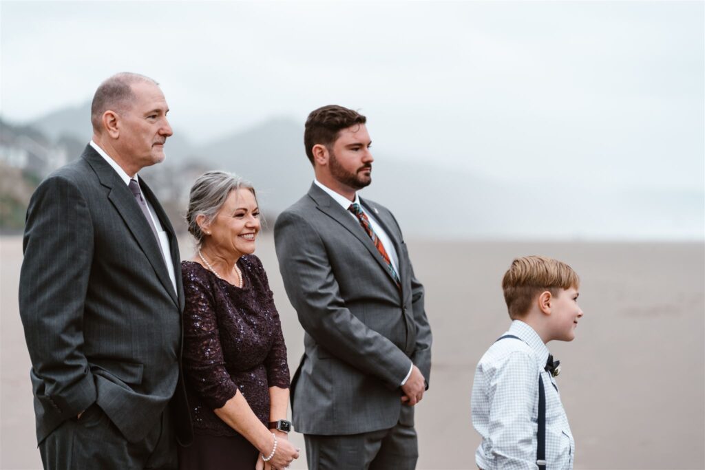 A shot of the grooms family reactions during the vow exchange on the gloomy beach. his mother, dressed for the occasion smiles excitedly as she witnesses their union during their pacific coast wedding 