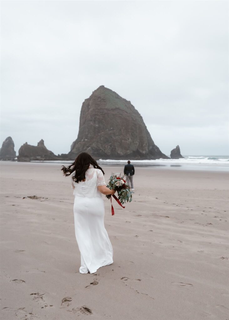 A bride walks out onto the empty, foggy beach to her groom. he faces away from her, waiting to be turned around for their first look during their pacific coast wedding 
