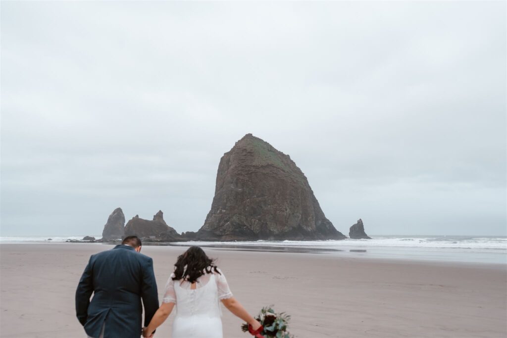 during their Pacific Coast wedding, a couple walks towards Haystack rock in their wedding attire.