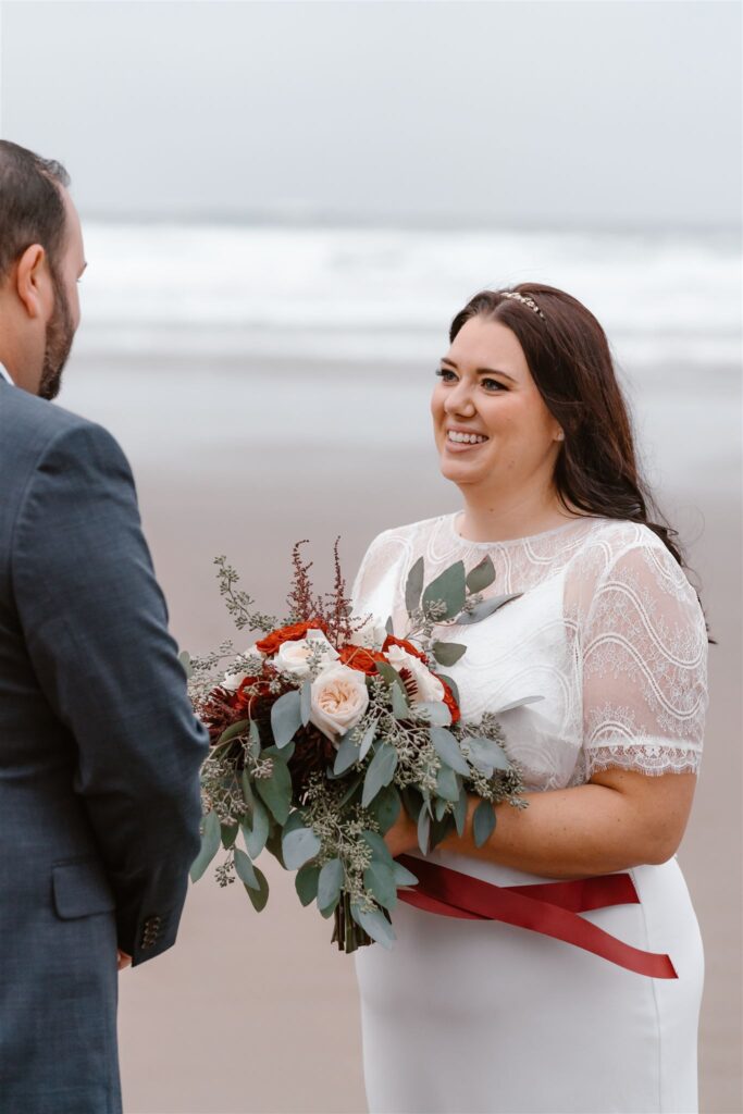 A bride clutching her bouquet of roses and greenery smiles lovingly at her groom. The wind blows the ribbon of her bouquet during their pacific coast wedding