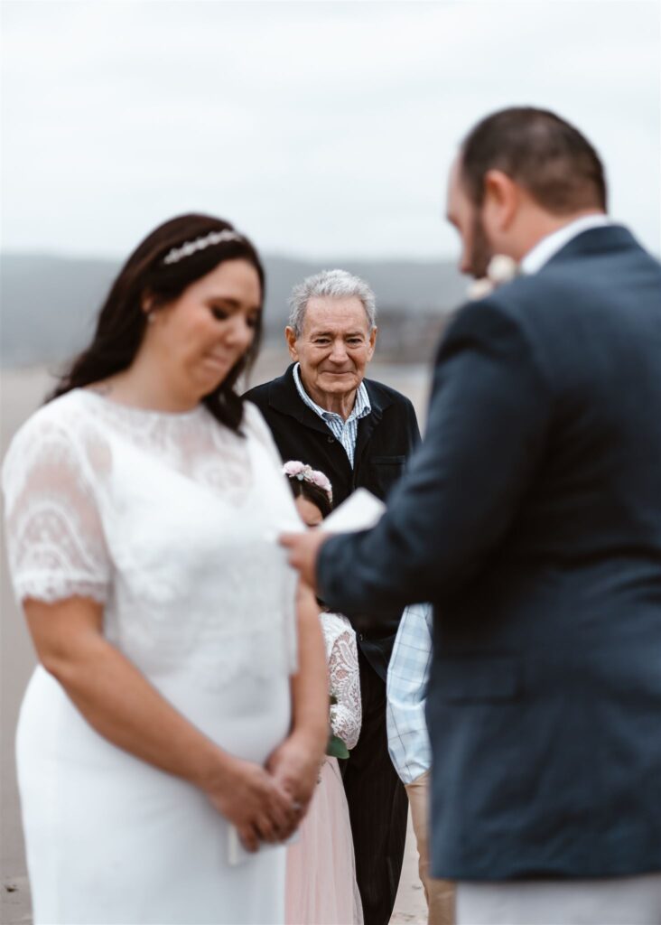 a perspective shot of the father of the bride crying joyfully. he is framed by the bride and groom. The groom is reading his vows to the bride during their pacific coast wedding.