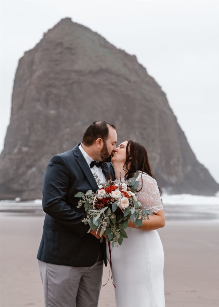 a bride and groom exchange their first kiss as married. they kiss in front of haystack rock as they celebrate their pacific coast wedding