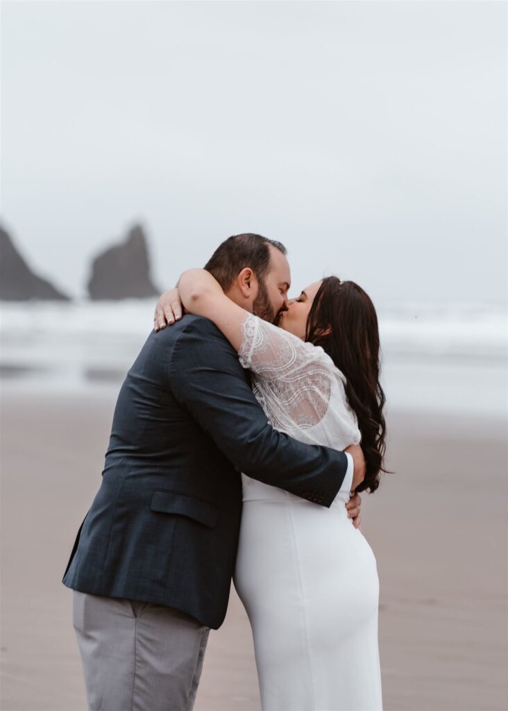a bride and groom exchange their first kiss as married. they kiss on the empty, moody beach as they celebrate their pacific coast wedding