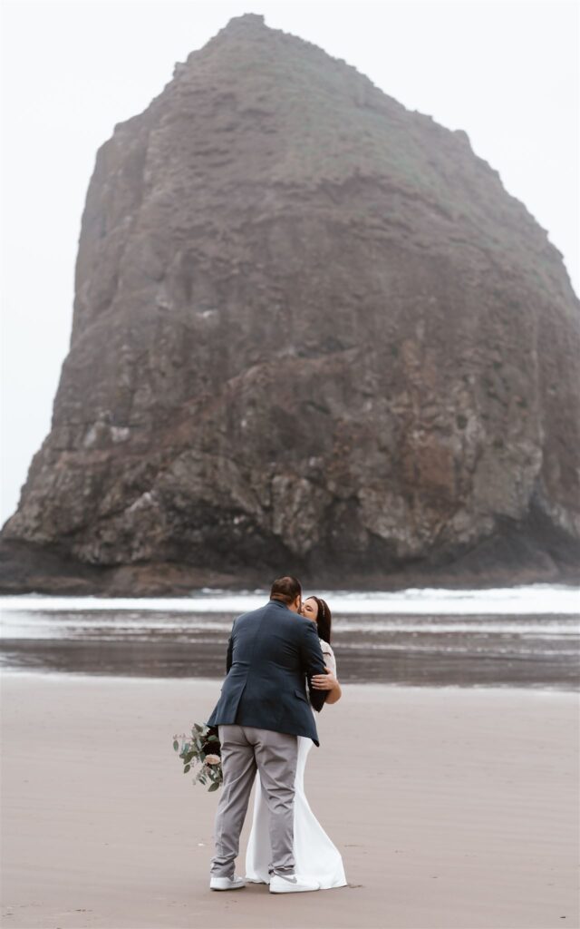 a couple in their wedding attire kiss in front of the iconic haystack rock on cannon, beach. The fog rolls in and creates a moody vibe for their Pacific Coast wedding. 