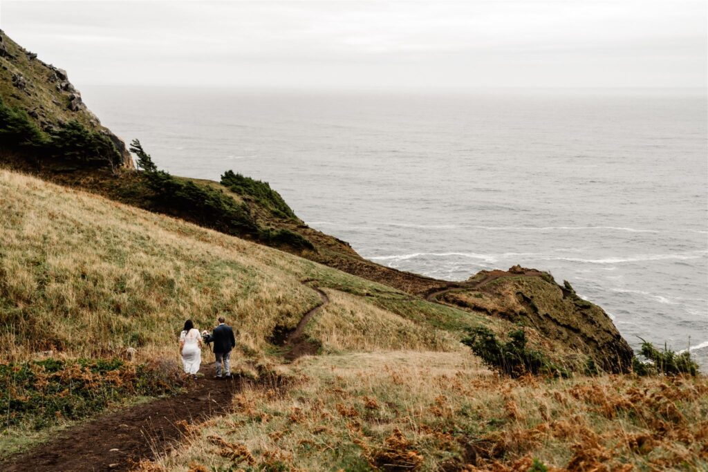 a wide landscape shot of a bride and groom explore a green trail that leads to an open cliffside during their pacific coast wedding 