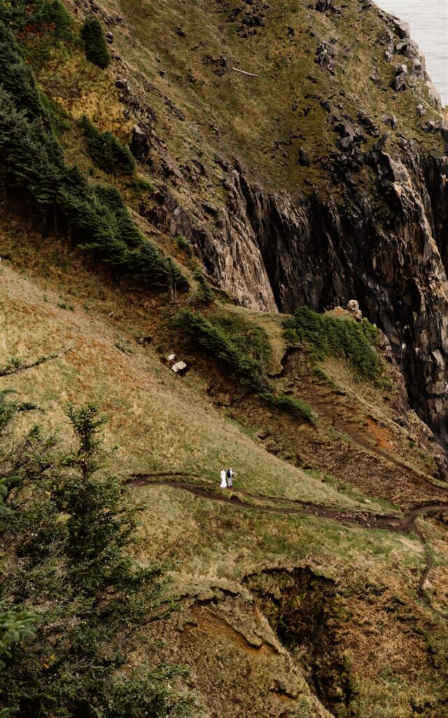 a large landscape shot of a couple in their wedding attire walking along a trail that is carved into a green cliff. The camera is far away, making the couple appear tiny as they explore during their Pacific Coast Wedding.