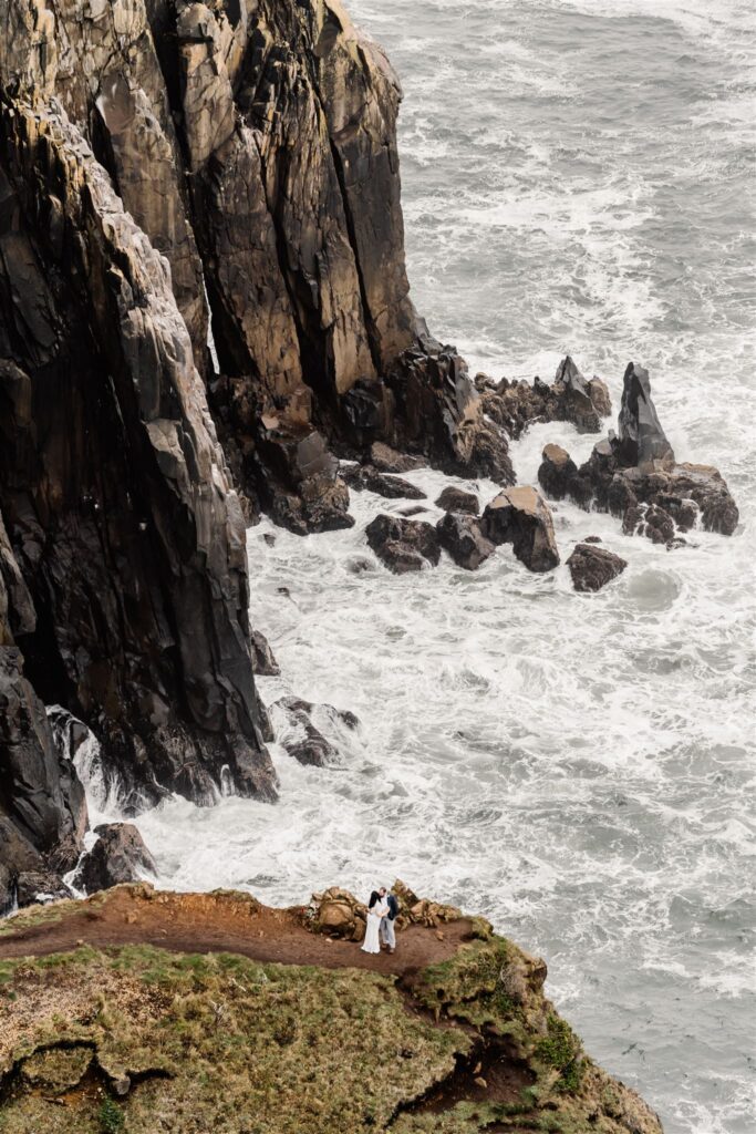 A shot to show off the landscape this couple chose to explore during their coastal elopement. They stand on the edge of a sea cliff with choppy ocean waves beneath them. They embrace in their wedding attire. 