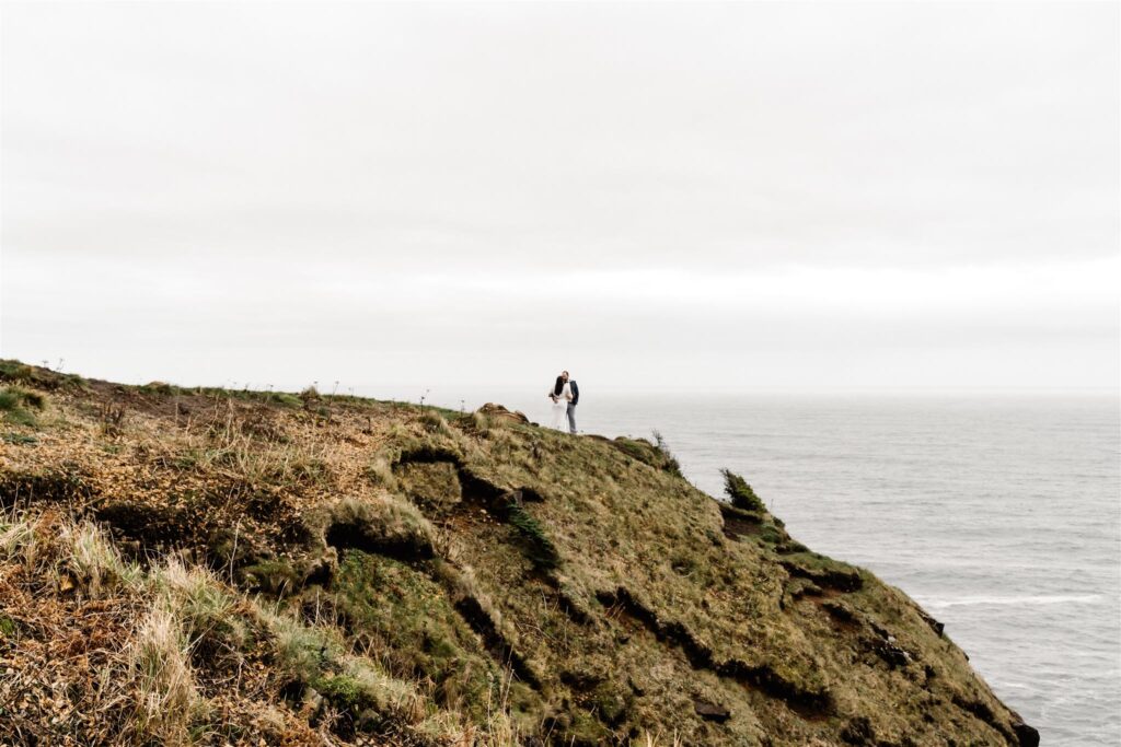 a bride and groom stand atop a green cliff. they kiss in their wedding attire. the cliff slopes down to the corner of the frame and the sky is gray and frames them.