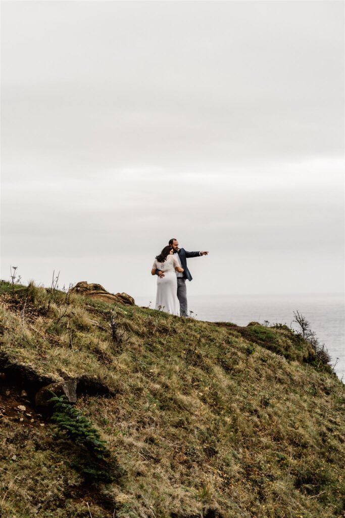 a couple in their wedding attire stands on a grassy cliff pointing out into the distance. They embrace as the groom points out into the distance. 