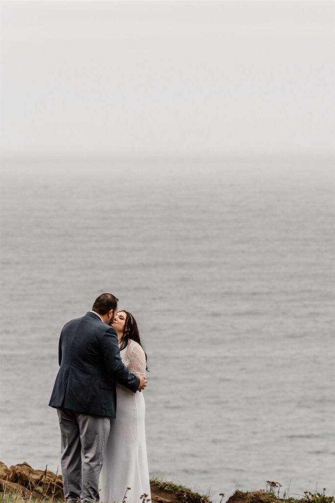 during their pacific coast wedding, a bride and groom kiss on the side of a cliff. the background is the icy gray ocean.
