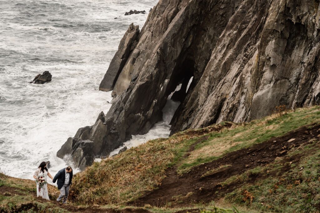 During their Pacific Coast Wedding, a couple walks a long the edge of a coastal cliff. The weather is moody and the ocean waves swirl beneath the basalt cliffs