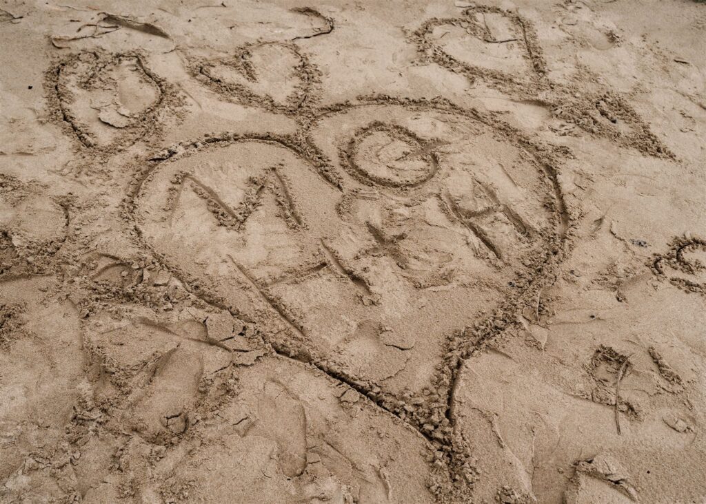 a detail shot of the initials of the bride and groom inside of a heart that is drawn in the sand to commemorate their pacific coast wedding 