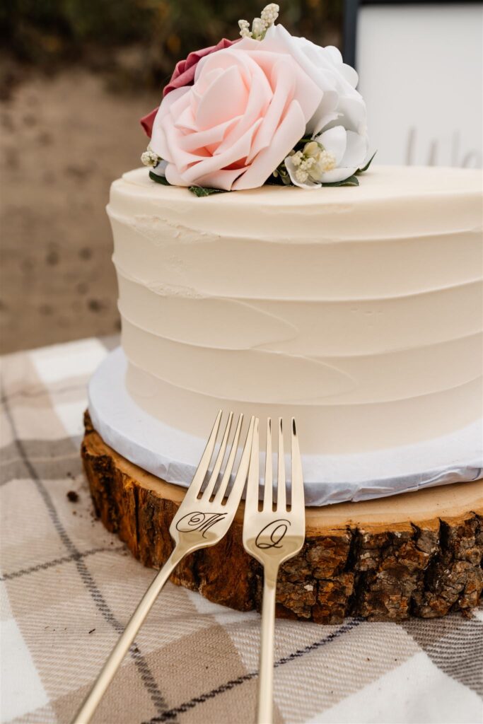 a detail shot of a small wedding cake topped with romantic, pink roses. it sits a top a round piece of oak. two golden forks lay near it with the intials of the bride and groom celebrating their pacific coast wedding