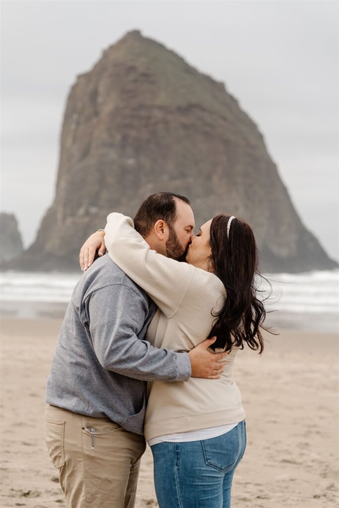 a bride and groom kiss in front of haystack rock. they are in comfy clothing and are celebrating their pacific coast wedding on the beach