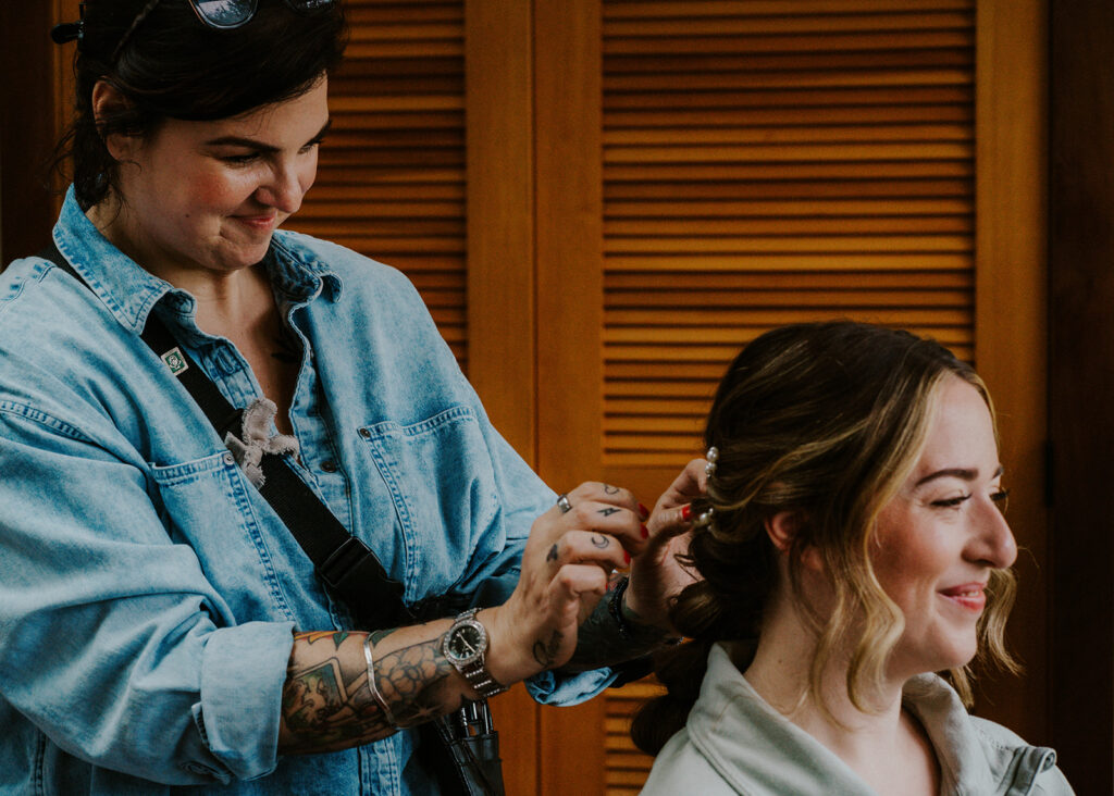 A makeup artist helps a smiling bride prepare for her forest wedding. She smiles as she tweaks her hair. 