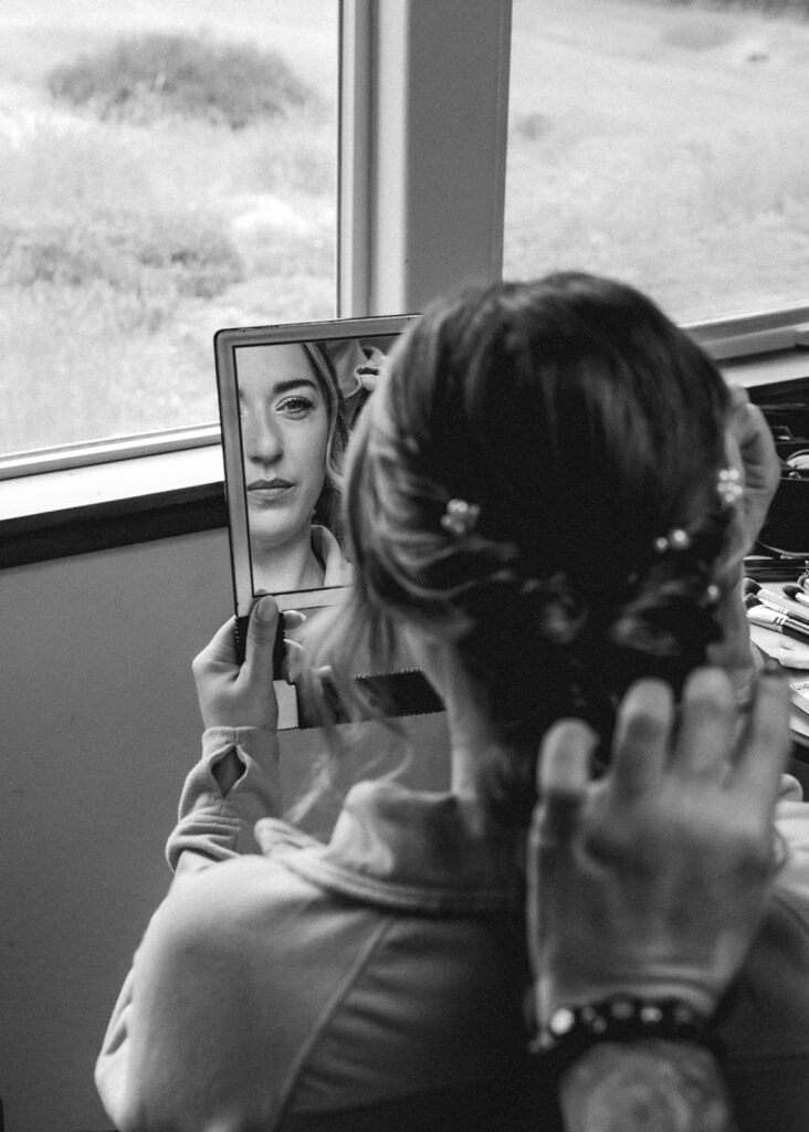 A vertical black and white photo of a bride getting ready using a handheld mirror. The focus is on the reflection of the bride, shot over her shoulder. 