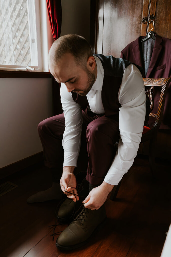 A groom in a deep maroon suit sits in a wooden chair. He laces up his hiking boots in preparation for his forest wedding. 