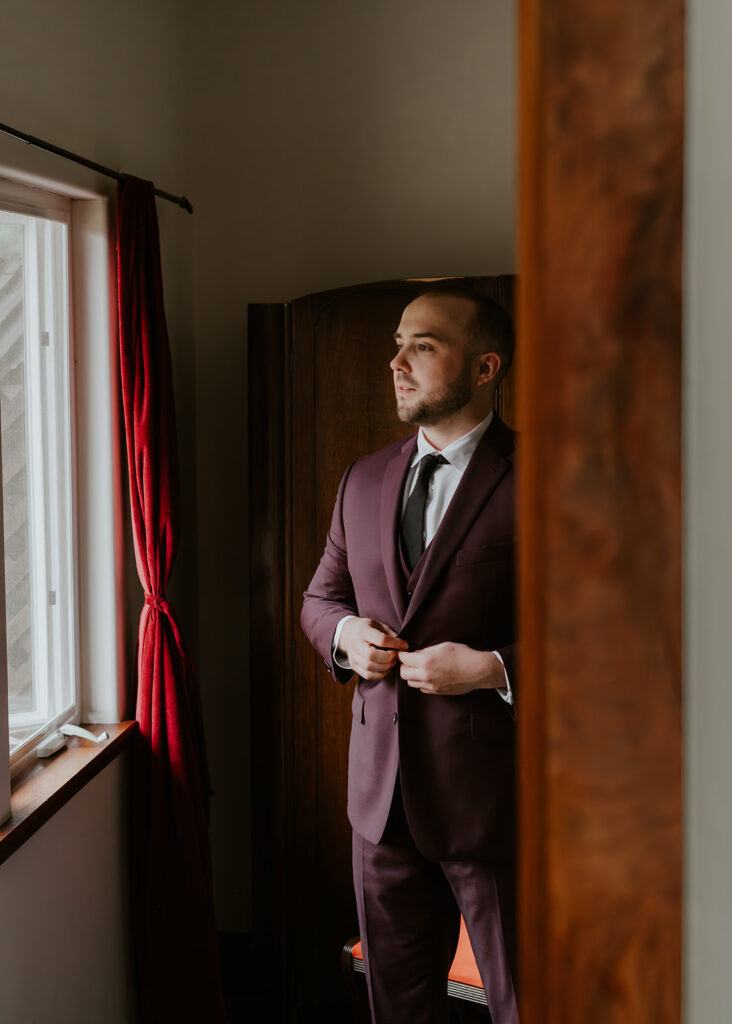 Framed by the doorway, we see a groom complete his forest wedding look. He buttons his deep maroon jacket as he gazes out the window.