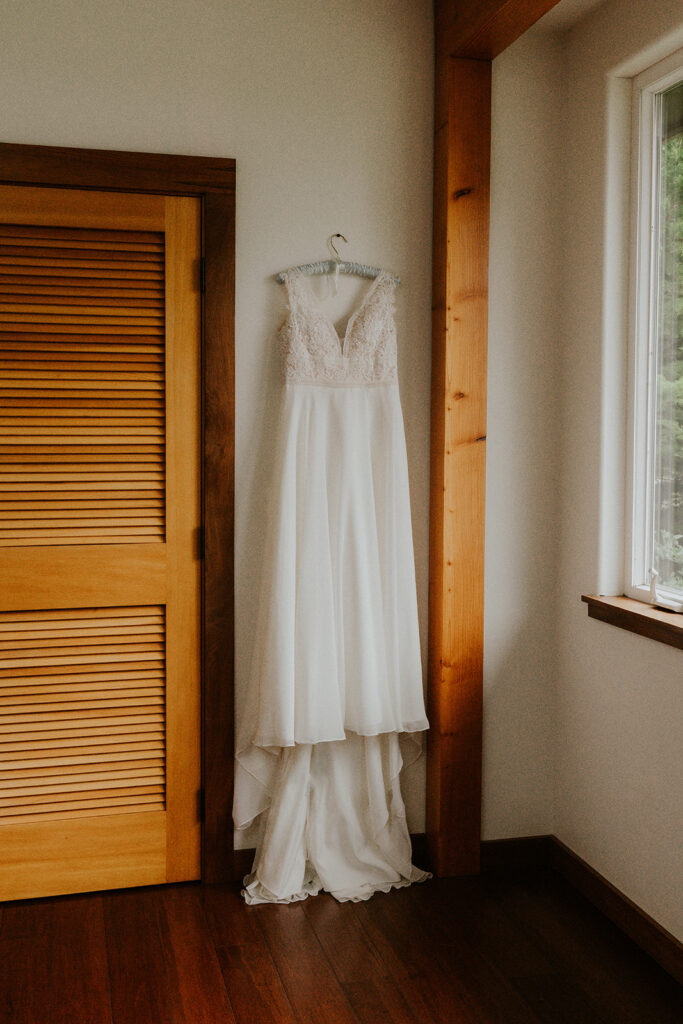 A vertical shot of the brides wedding dress hanging on the wall. A window provides soft light. 