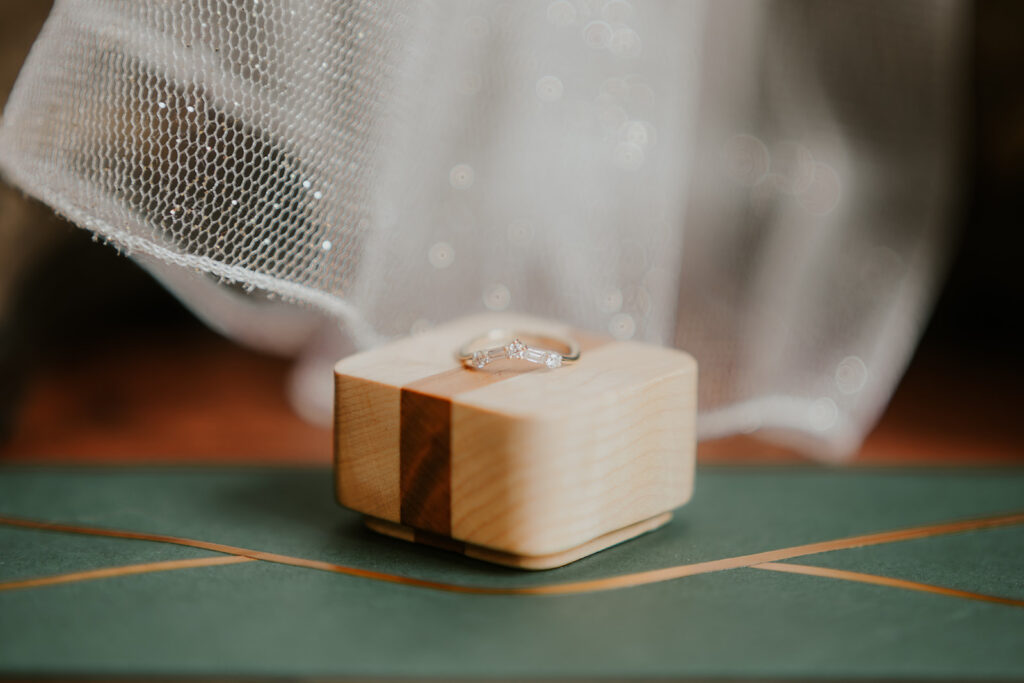 A shot of some of the details for this forest wedding. A wedding ring sitting atop a wooden box. The box rests on an emerald green envelop. In the background, the sparkles of a veil can be seen.