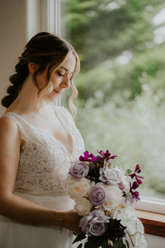 A bride stands in her dress by a window, letting in soft light. she gazes down at her bouquet of purple and white flowers.  