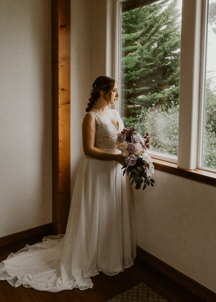 A bride stands gazing out the window to a garden in her wedding dress. The lighting is soft and the moment feels quiet as she waits to meet her groom.