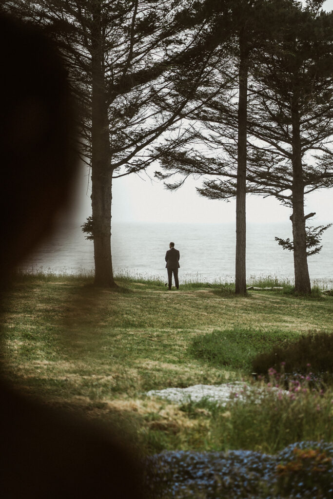 Shot over our brides shoulder, her head and shoulder framing the view of her groom standing by the ocean cliffs, waiting to see her. A spray of bright flowers from the garden add pops of color to the scene. 