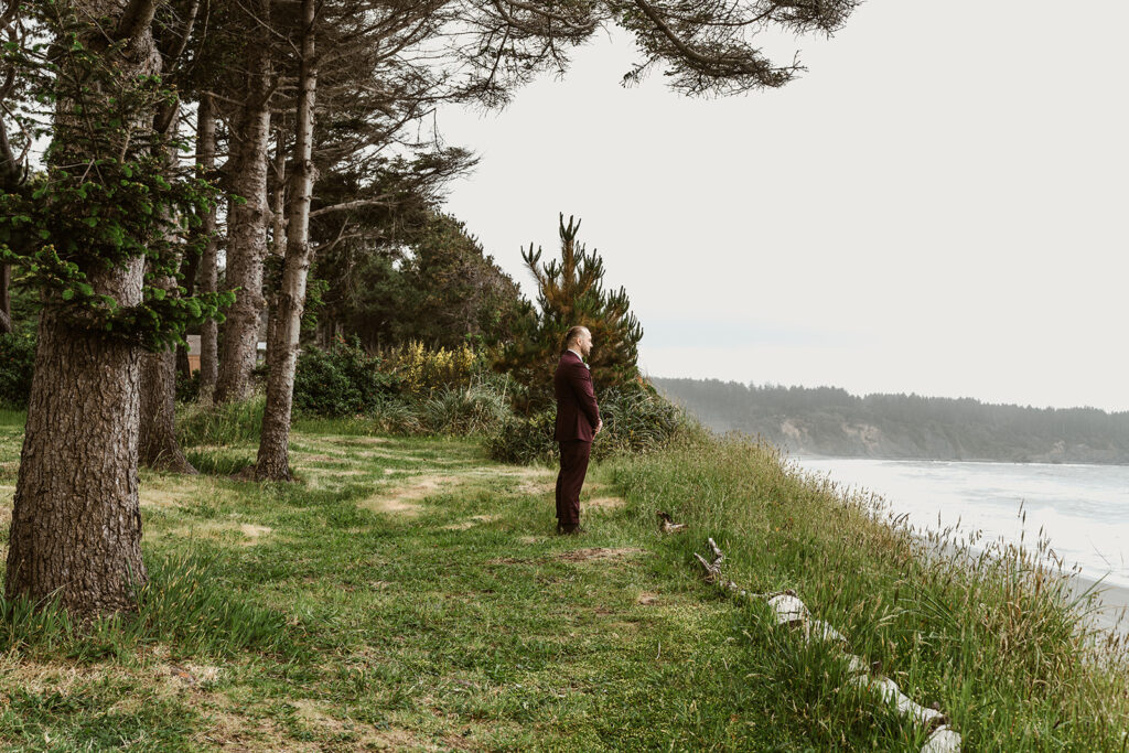 A groom waits to see his bride. He stands on a grassy bluff in his maroon suit 