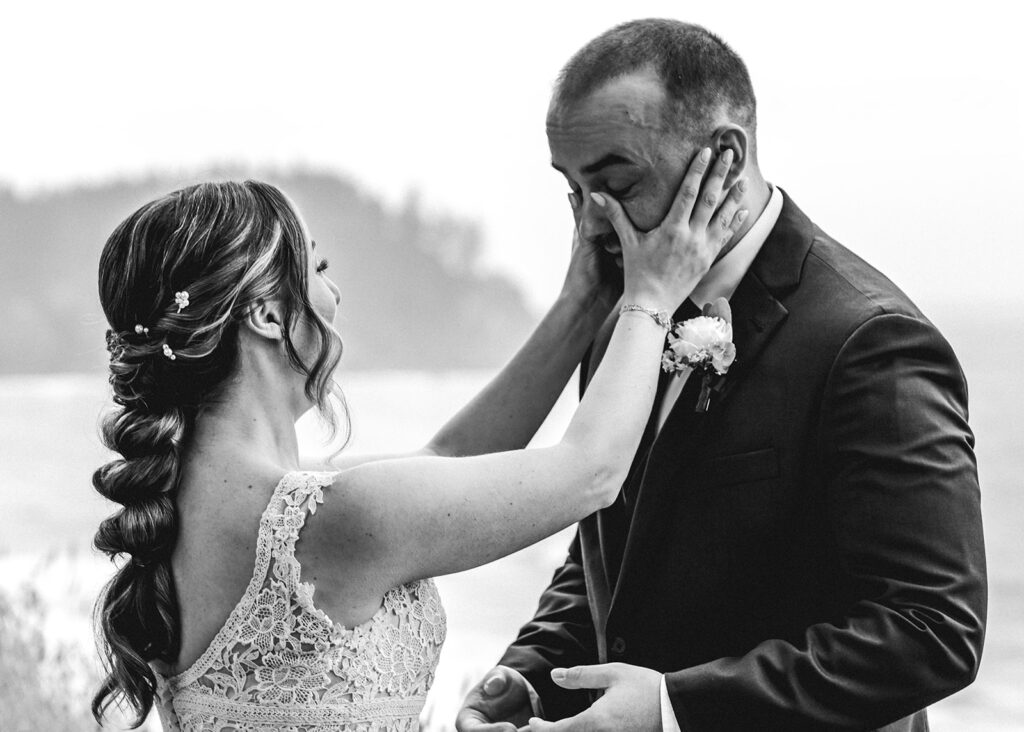 A black and white photo as a bride wipes the tear of her groom after he sees her for the first time in her wedding attire before their forest wedding