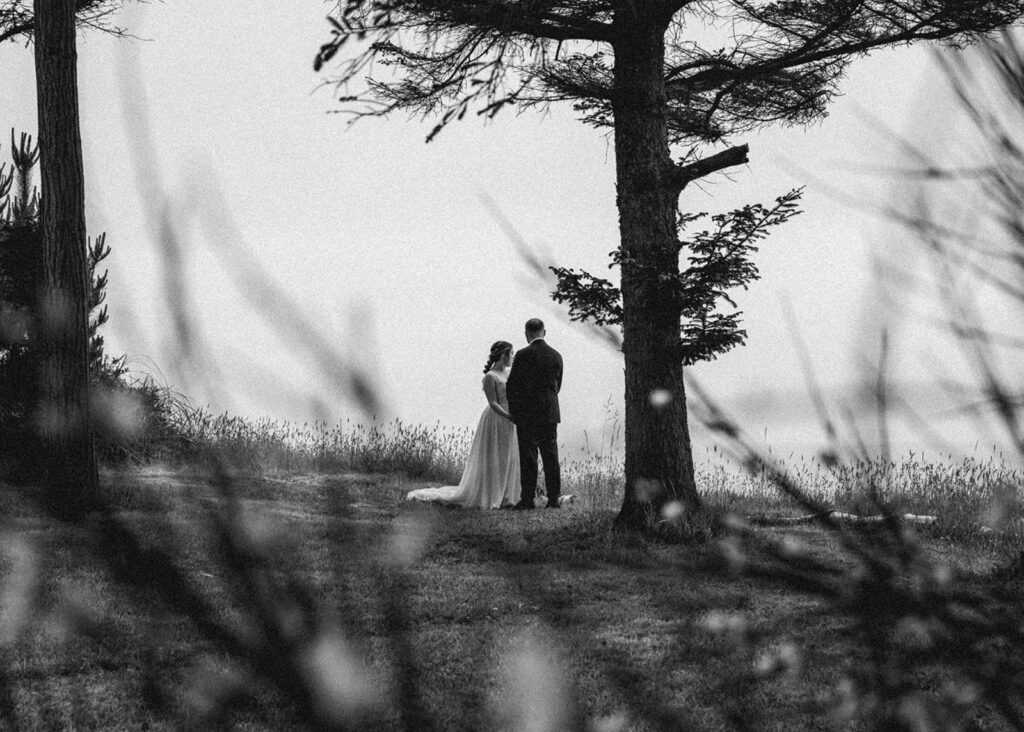 A black and white photo of a bride and groom in their wedding attire. They stand beside a cliff and are framed by the flowers around them. 