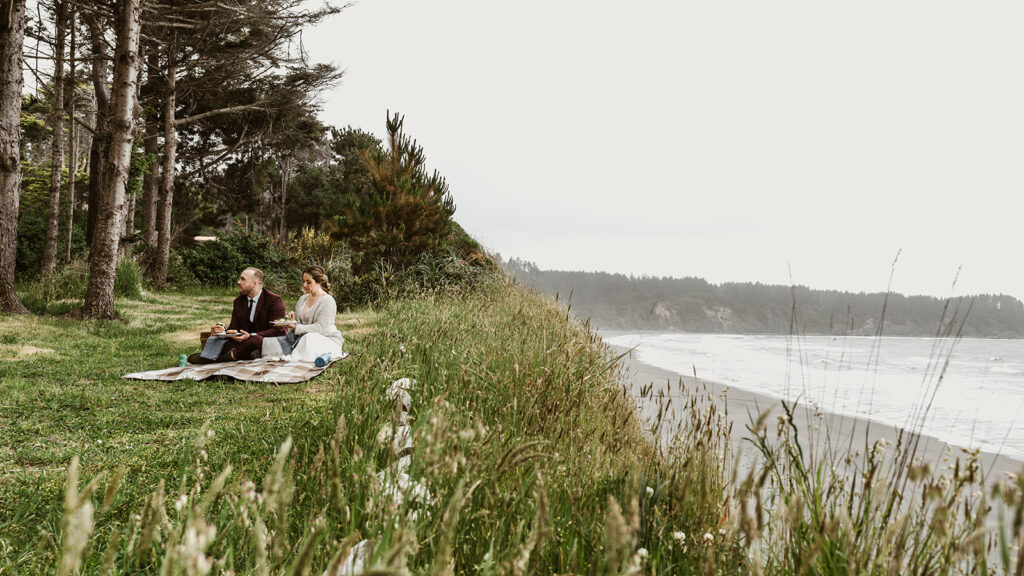 during a picnic, a bride and groom eat  sitting on a blanket. they are sitting cliffside with the ocean below them.