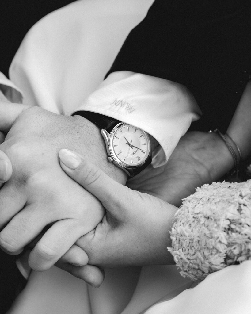 a detailed, black and white shot of the bride and groom holding hands. The groom wears a sleek watched just gifted to him by his bride for their forest wedding. 