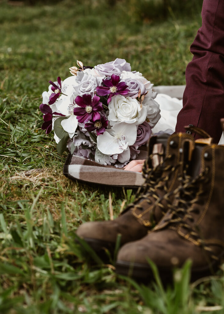 A vertical, detail shot of the brides purple and white bouquet laying on the ground, framed by green grass and hiking boots